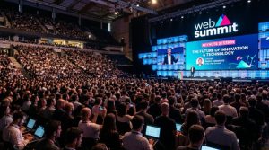 Packed audience at a Web Summit keynote session with speaker on stage and large LED screen in background
